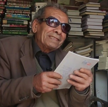 Ahmad Zereeni, founder of Irbid’s iconic book booth, smiles while holding a book, surrounded by shelves of stacked titles.u2028(Photo by Hussein Zereeni)